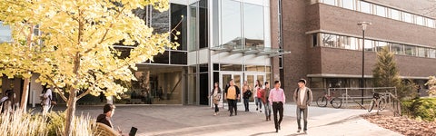 A group of students walking in front of Hagey Hall building, surrounded by fall trees.